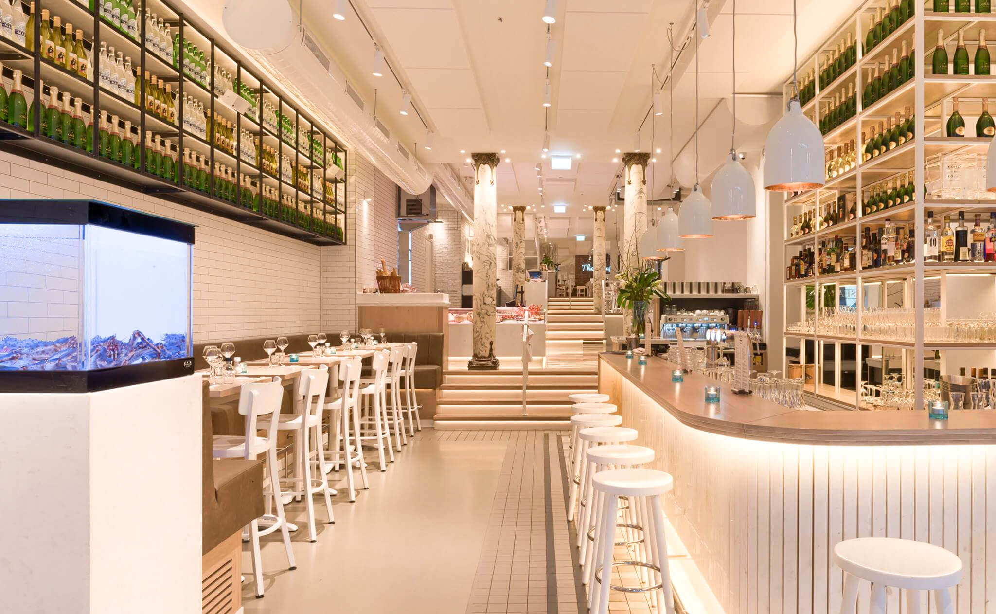 Světlá restaurace se zaobleným barem, bílými barovými židlemi a policemi s lahvemi vína.
